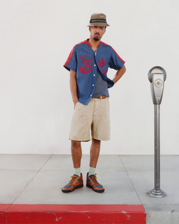 A man in an Aimé Baseball Jersey, khaki shorts, and hat stands by a parking meter on a red-curbed sidewalk.