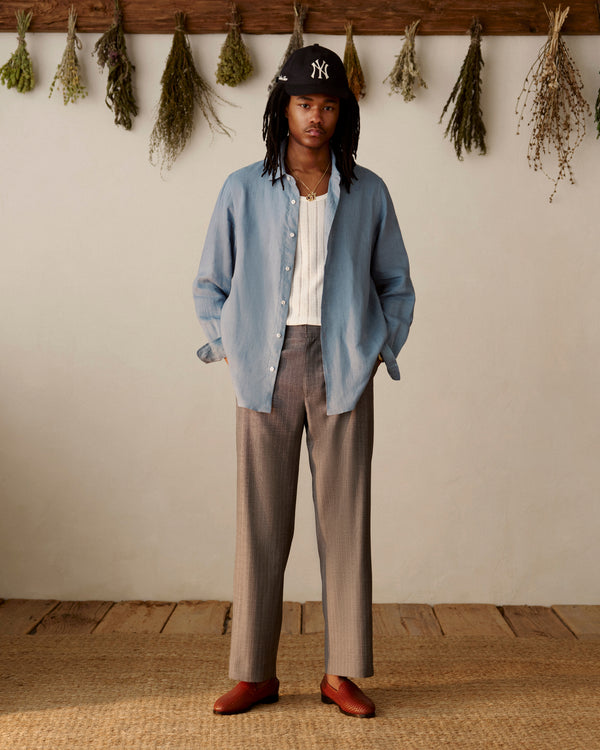 A person in a light blue shirt and Pinstripe Elasticated Waist Trouser stands indoors on a woven rug with dried herbs behind.