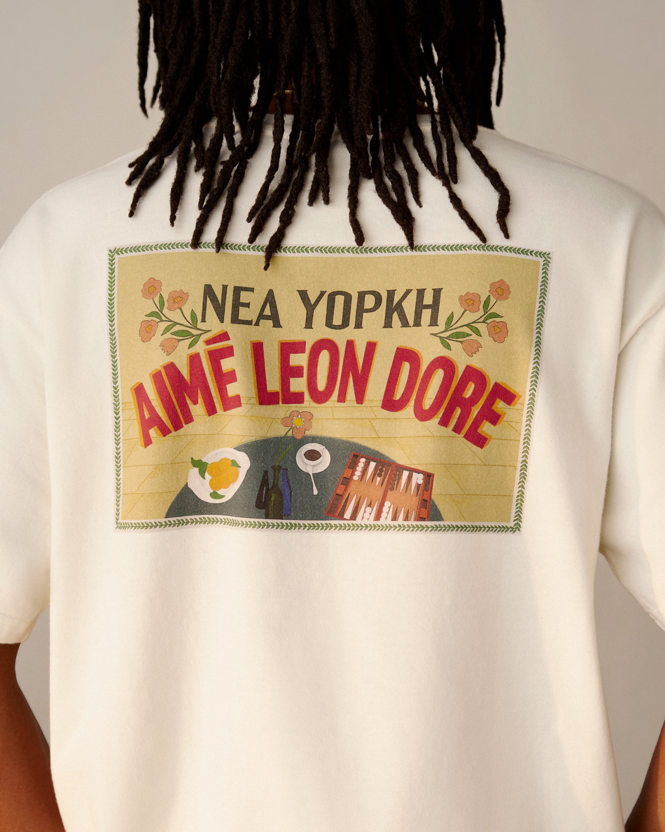 Person wearing a white Aimé Postcard Tee with food and building graphics on the back.