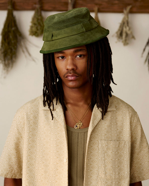 A person with long dreadlocks wears a Perforated Suede Bucket Hat and necklaces, standing indoors with herbs behind.