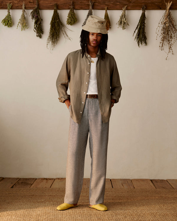 Someone in a Long-Sleeve Linen Shirt and hat stands indoors, with dried herbs hanging on the wall behind them.