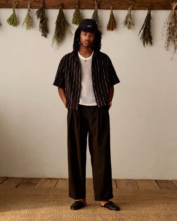 A person in loose black pants, a striped shirt, a cap, and Leather Raffia Mules stands indoors near dried herbs.