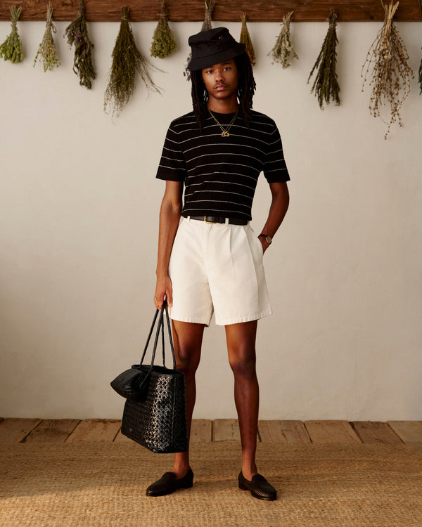 A person in a Merino Wool Eyelet Tee, white shorts, black hat, and loafers stands indoors holding a large black bag.