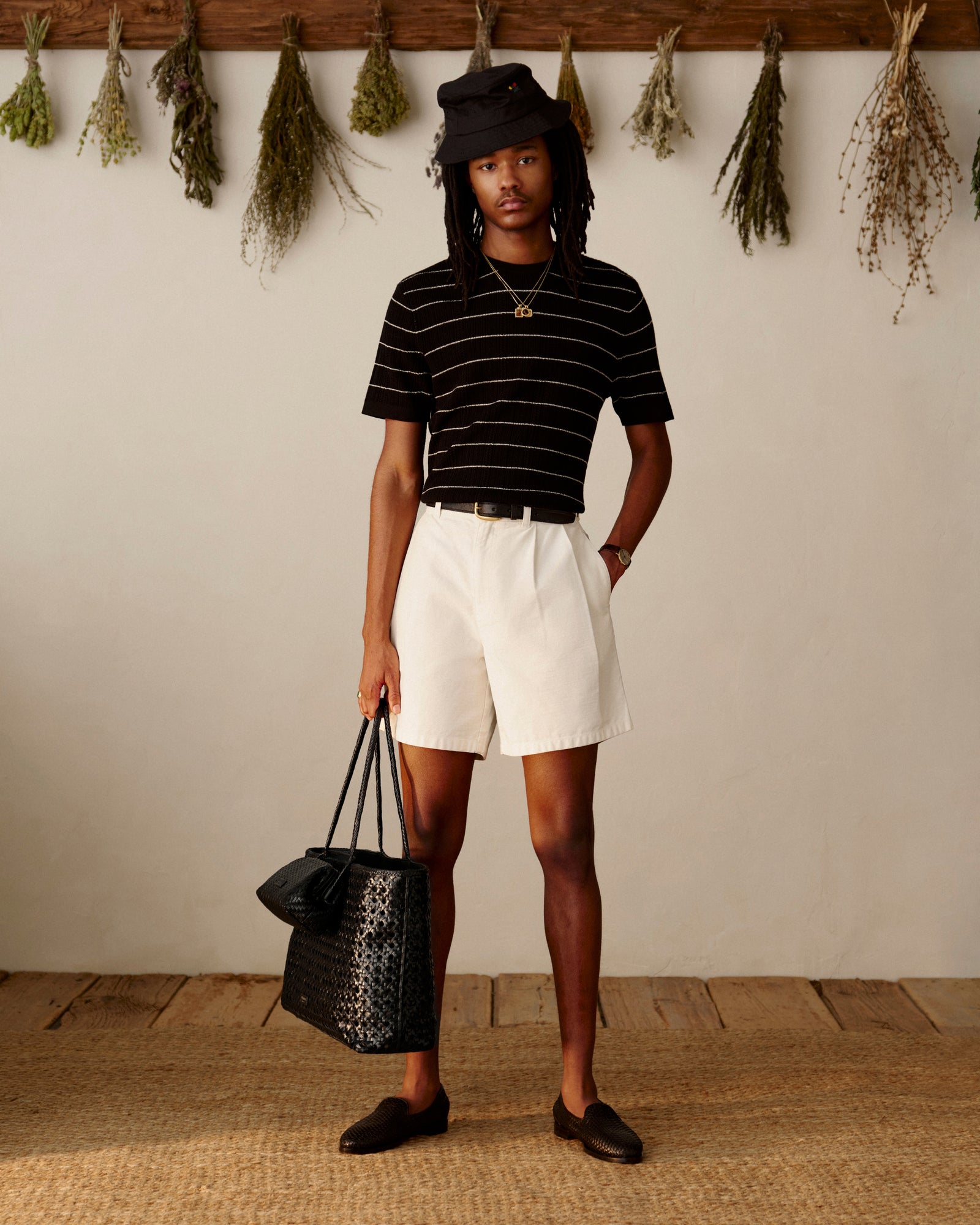 Person wears striped black shirt, white shorts, black Leather Raffia Loafers, and holds a black bag indoors by hanging herbs.
