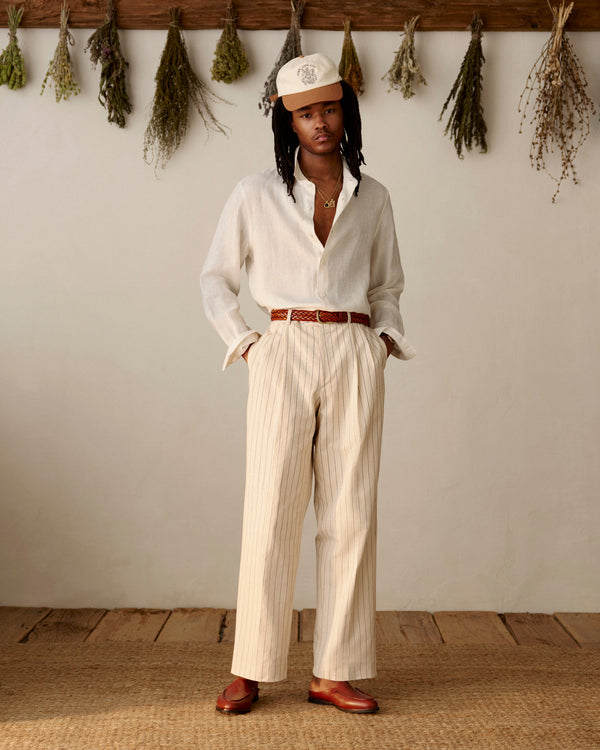 Wearing the Saint George Logo Hat, a person in light clothes and loafers stands indoors by dried herbs on the wall.