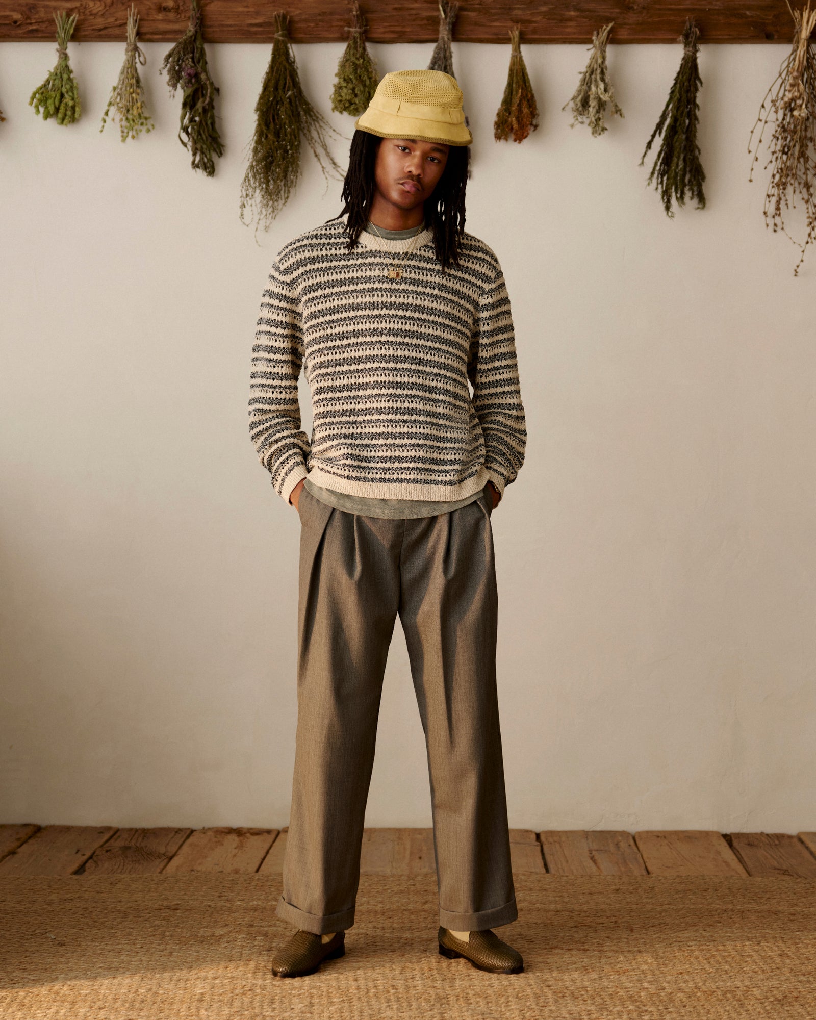 Person in striped sweater, yellow bucket hat, loafers, and Lightweight Cuffed Trouser stands indoors by hanging dried herbs.