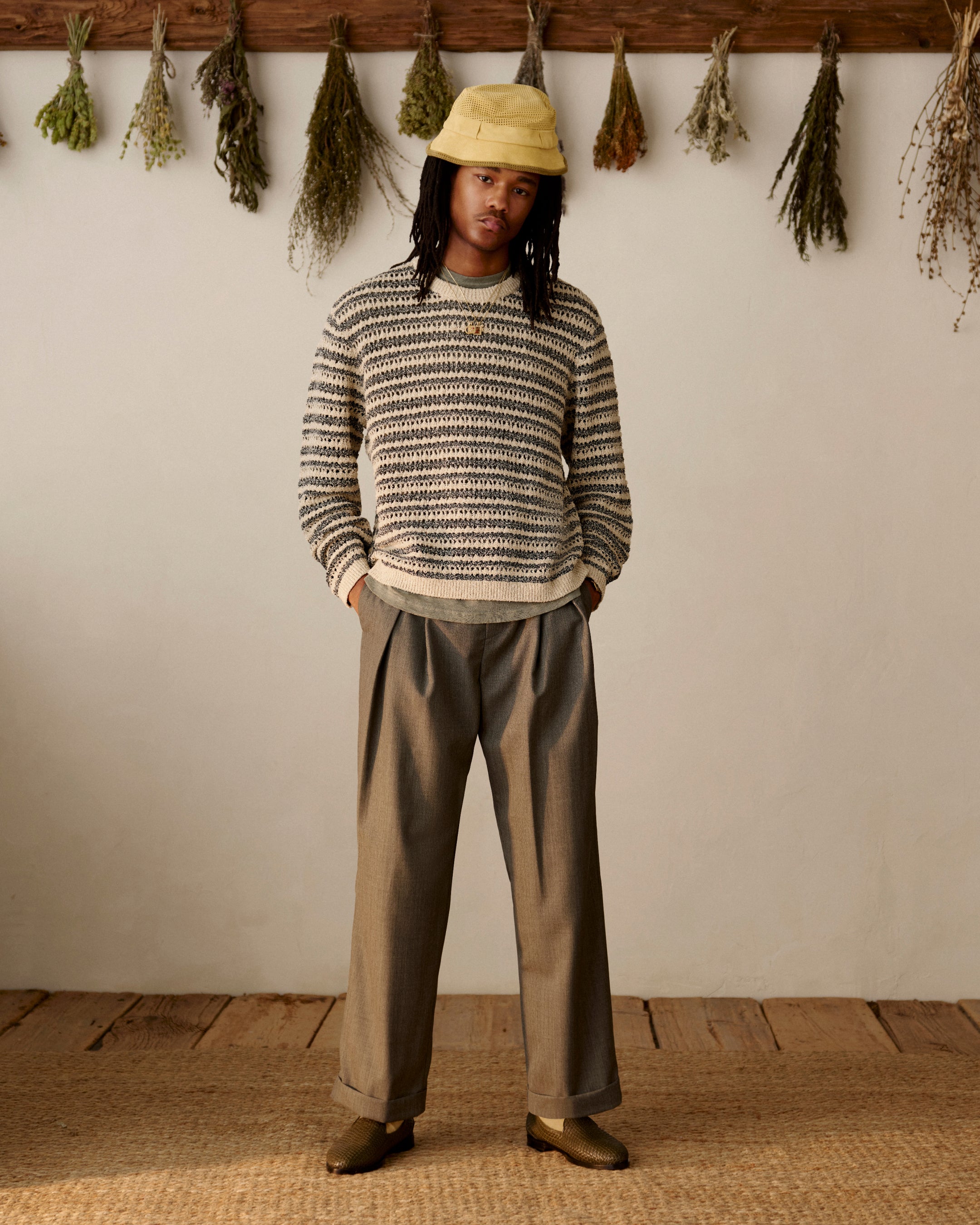 Person in striped sweater, yellow bucket hat, loafers, and Lightweight Cuffed Trouser stands indoors by hanging dried herbs.
