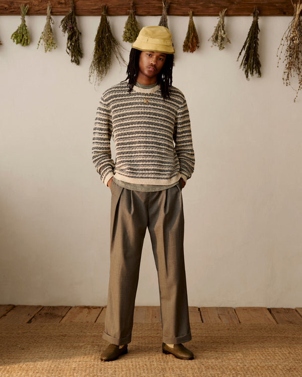 Person wears Leather Raffia Loafer, striped sweater, wide brown pants, and yellow hat indoors with dried herbs behind.