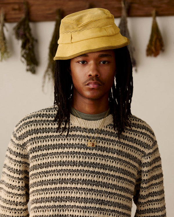 Person with long hair wearing a Perforated Suede Bucket Hat and striped sweater, indoors with dried herbs in the background.