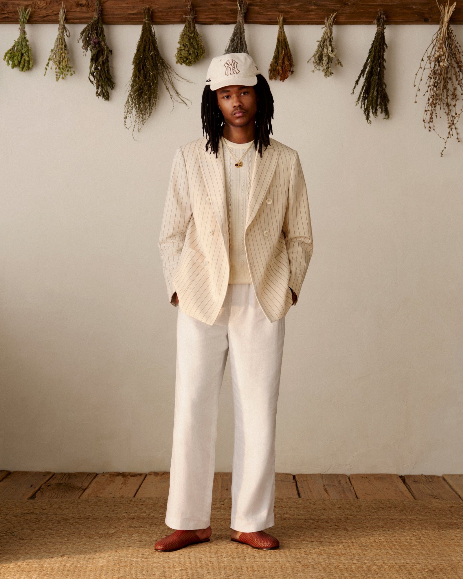 Someone wears a Double-Breasted Pinstripe Suit Jacket and white cap indoors with dried herbs hanging behind them.
