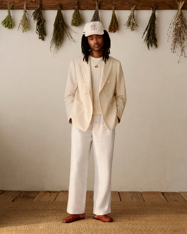 A person in Linen Leisure Pants, white shirt, and cap stands indoors on a wooden floor with dried herbs hanging behind.