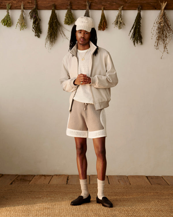 A person in Knit Court Shorts and a beige jacket stands indoors with dried herbs hanging on the wall behind them.