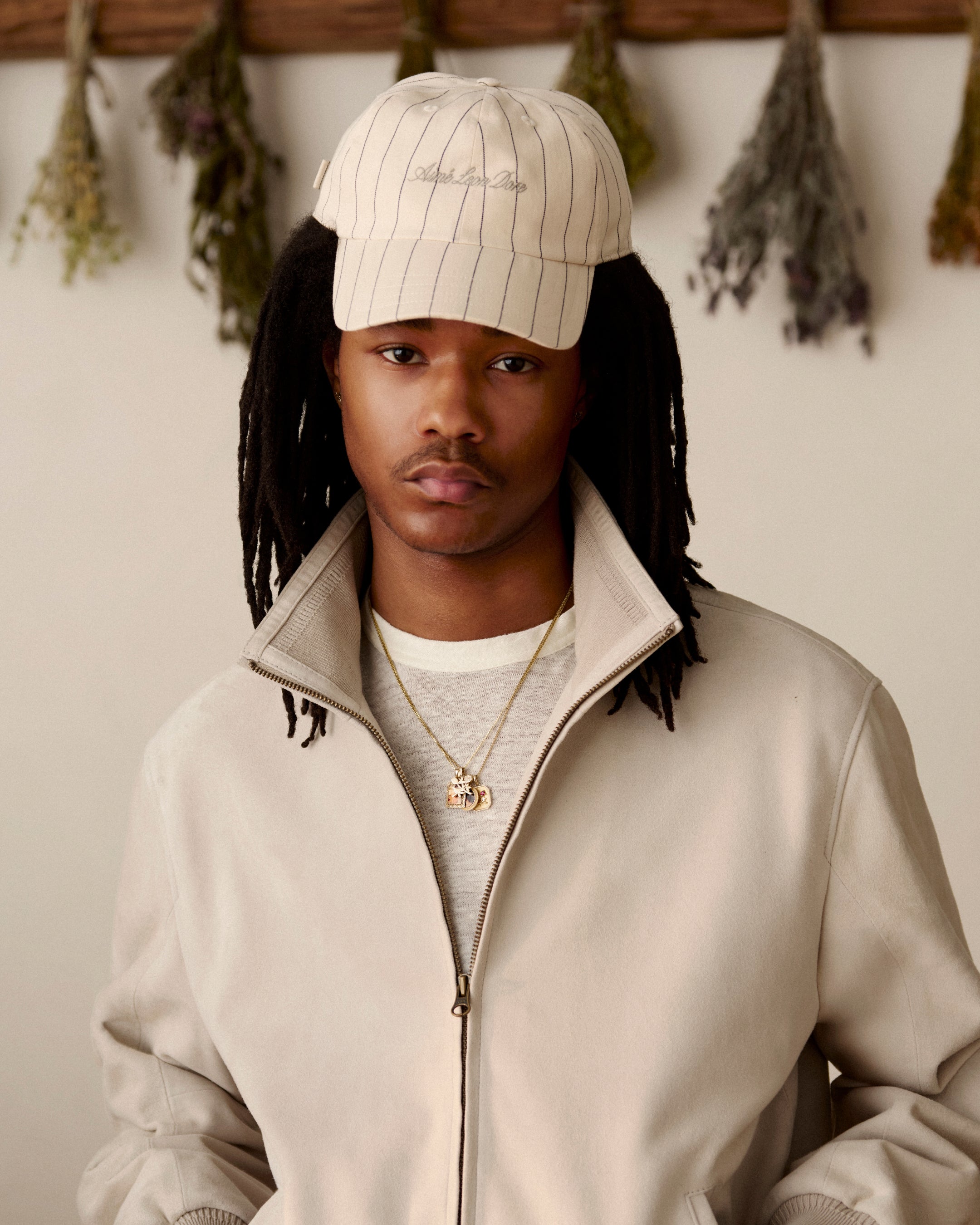 A man in a beige jacket and a Pinstripe Suiting Hat stands indoors with dried herbs hanging behind him.