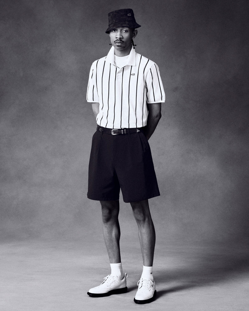 Man wears ALD Golf Polo, black shorts, bucket hat, and white shoes while standing against a plain backdrop.