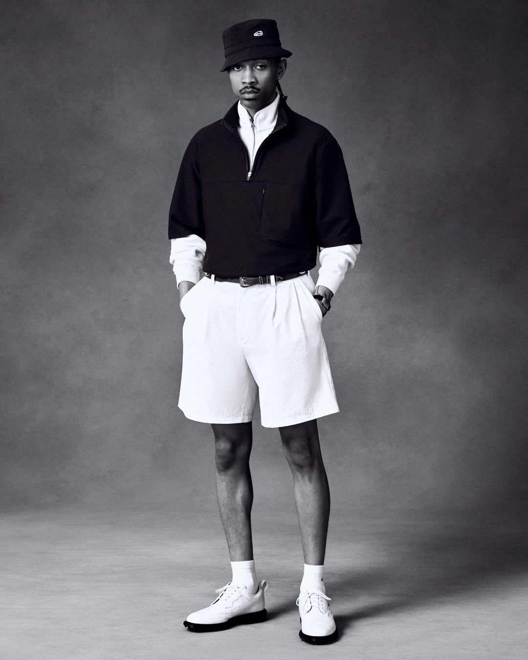 A man in a bucket hat, zip-up pullover, white shirt, shorts, and ALD / Footjoy Croc-Embossed Marquis poses simply.