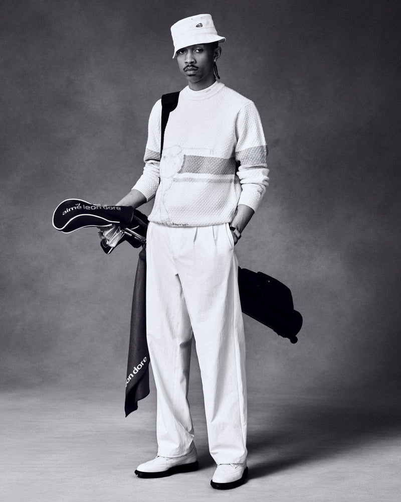 Black-and-white photo of a man in a bucket hat holding golf clubs, wearing the ALD Golf Clubhouse Sweater.