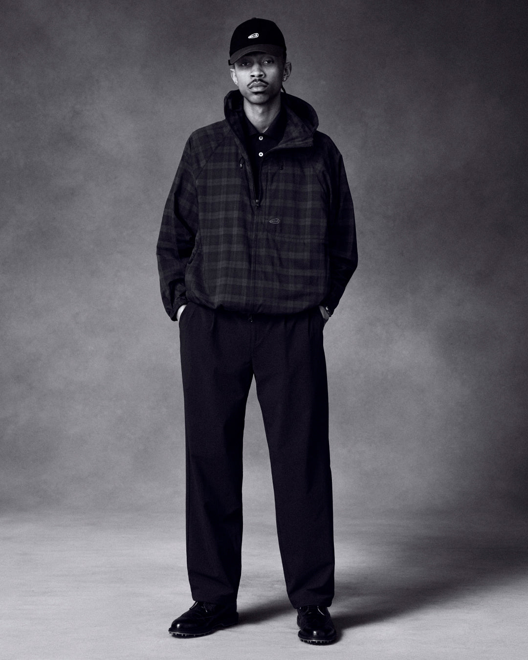 A man stands with hands in pockets, wearing a plaid jacket, wide-leg pants, ALD Golf Logo Hat, and black shoes.
