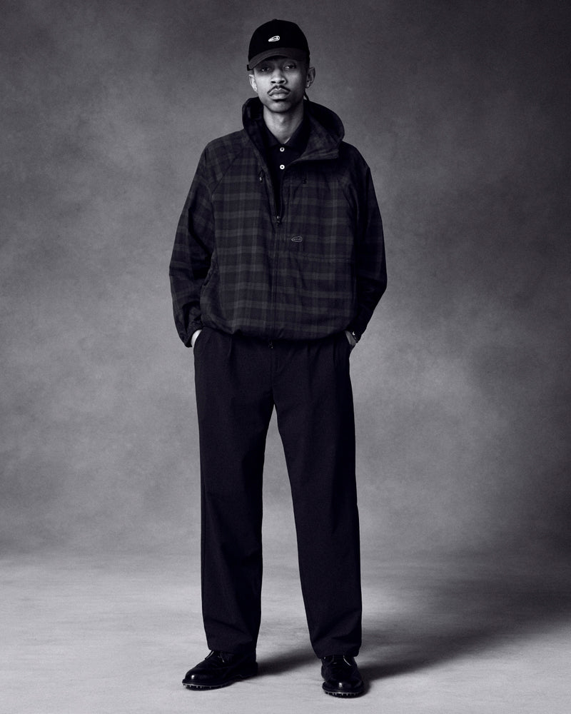 A man stands with hands in pockets, wearing a plaid jacket, wide-leg pants, ALD Golf Logo Hat, and black shoes.