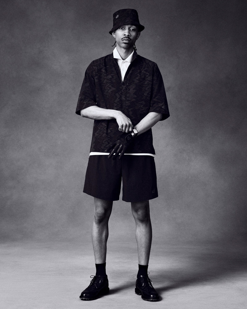 A man in ALD Golf Performance Short, bucket hat, dark shirt, gloves, and dress shoes stands against a plain background.