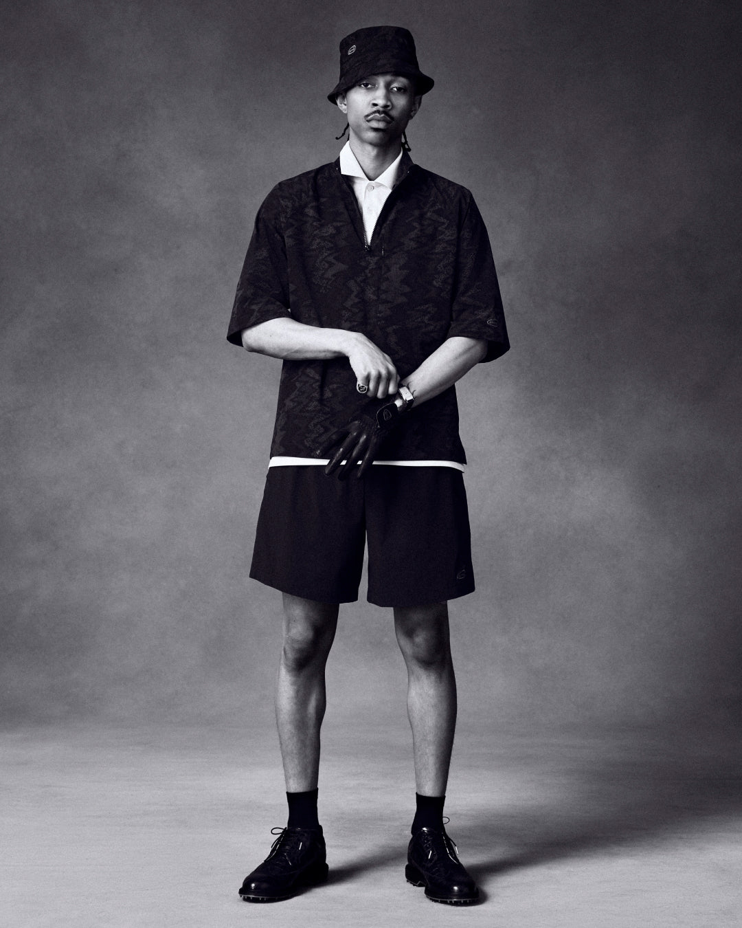 A man in a bucket hat, dark shirt, and shorts showcases the ALD / Footjoy Croc-Embossed Premiere Series Marquis.