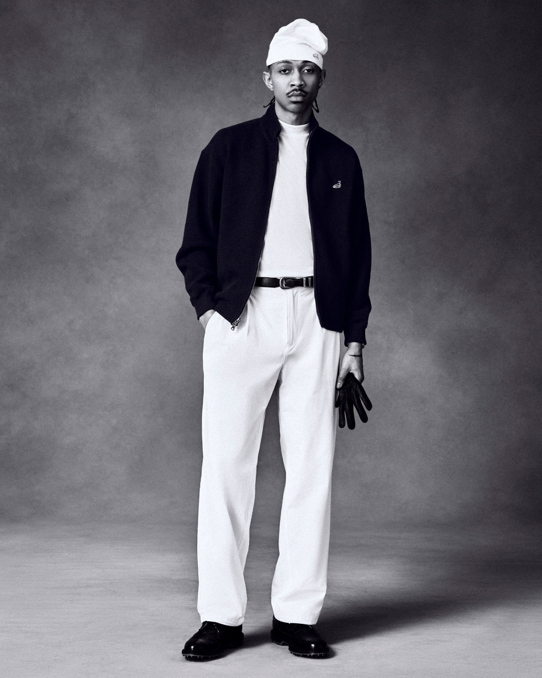 A man in an ALD Golf Piqué Full Zip Sweater, white pants, and cap holds black gloves against a dark background.