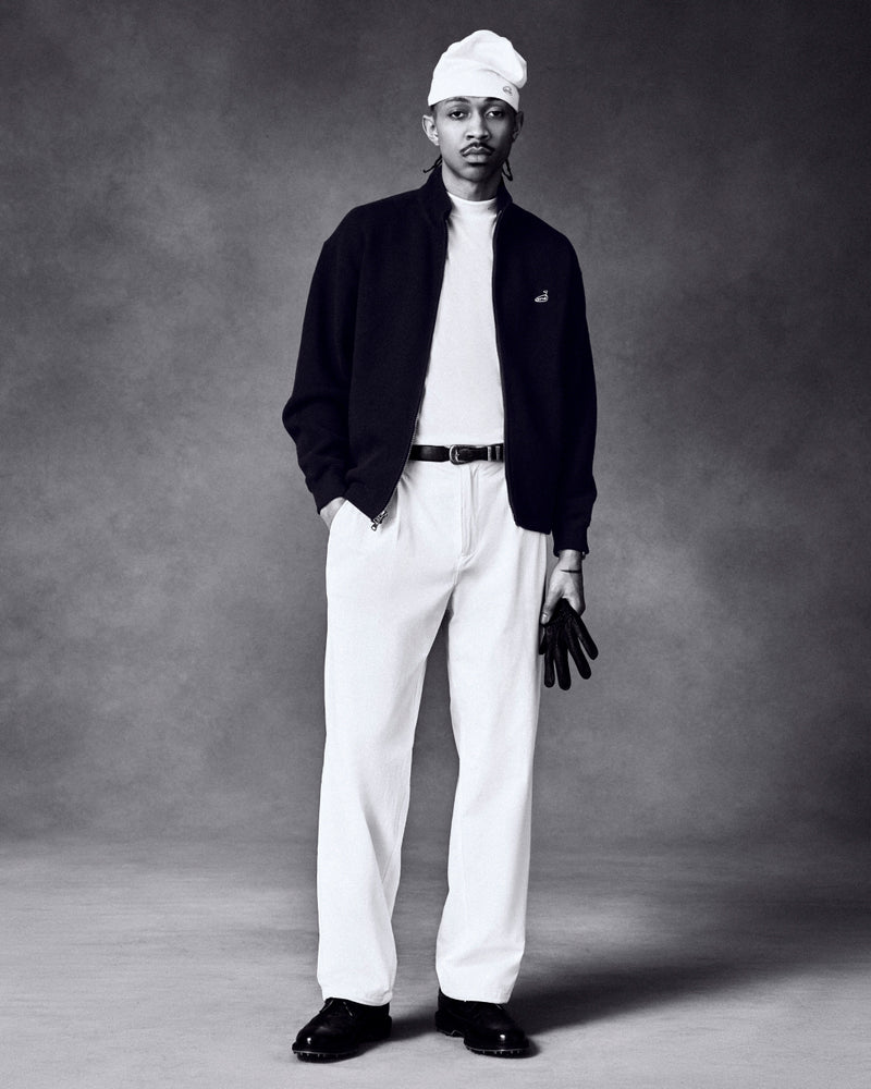 A man in an ALD Golf Piqué Full Zip Sweater, white pants, and cap holds black gloves against a dark background.