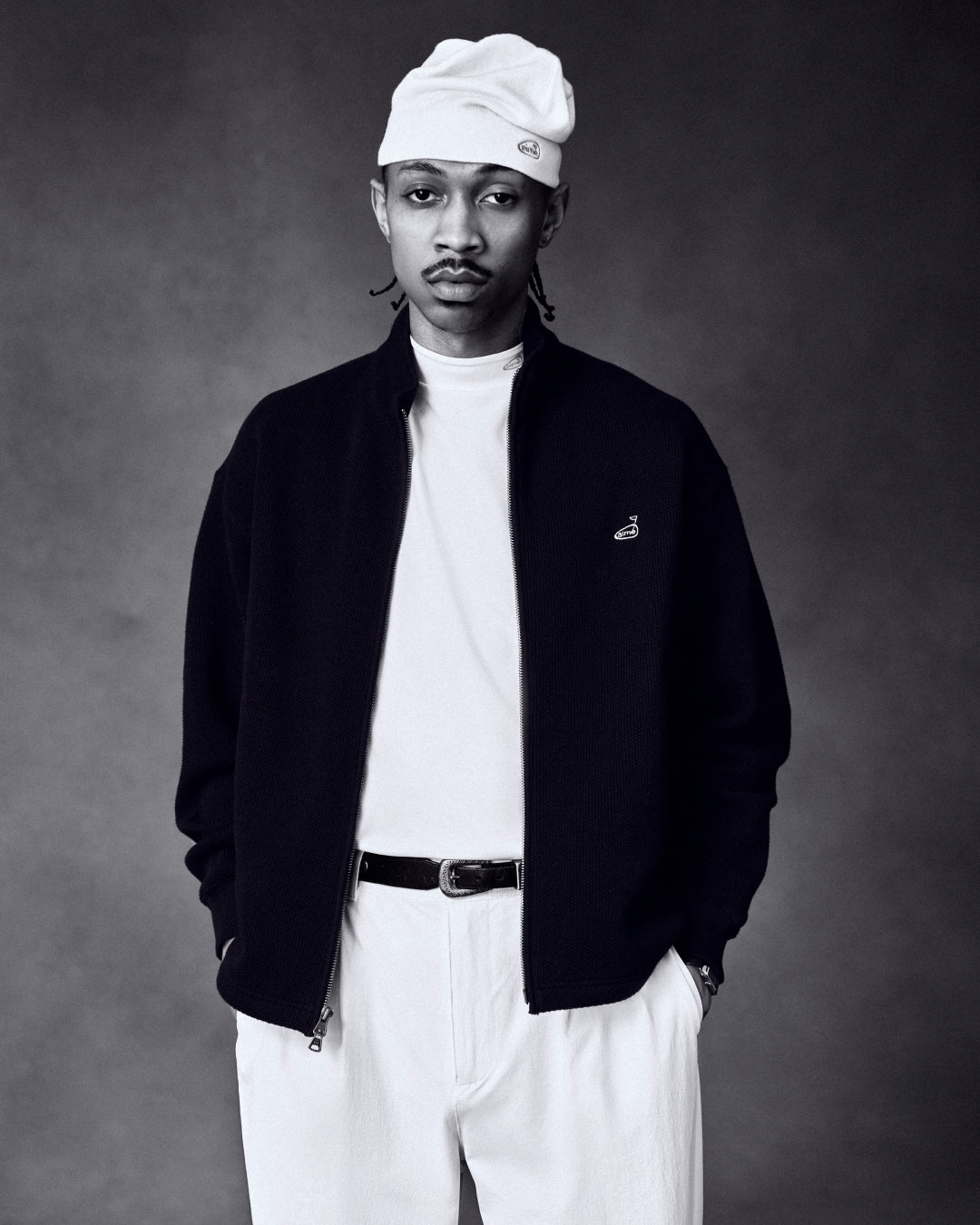 Man in ALD Golf Performance Tee, light beanie, dark jacket, and white pants, hands in pockets against dark background.