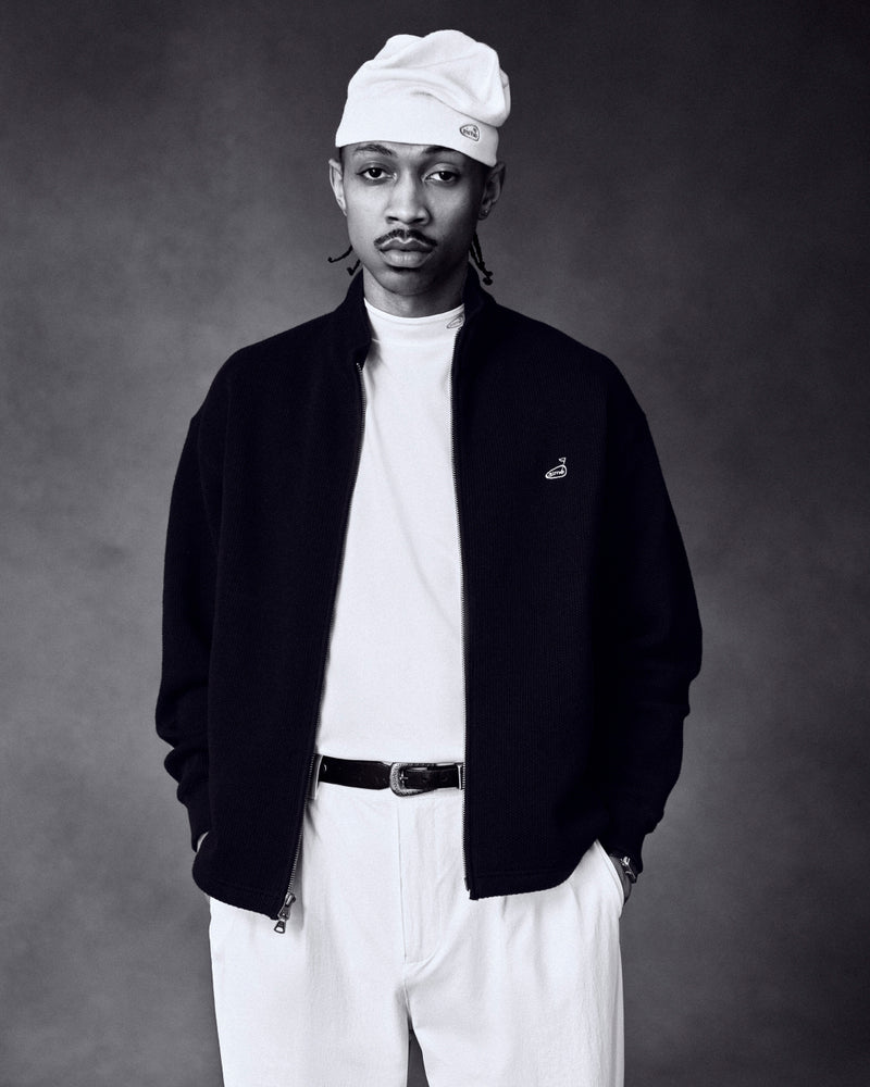 A man in an ALD Golf Skull Cap, white shirt, black jacket, and white pants stands with hands in pockets.