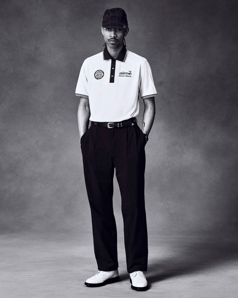 A man in a white polo, dark pants, ALD Golf Performance Sport Hat, and white shoes stands against a gray backdrop.
