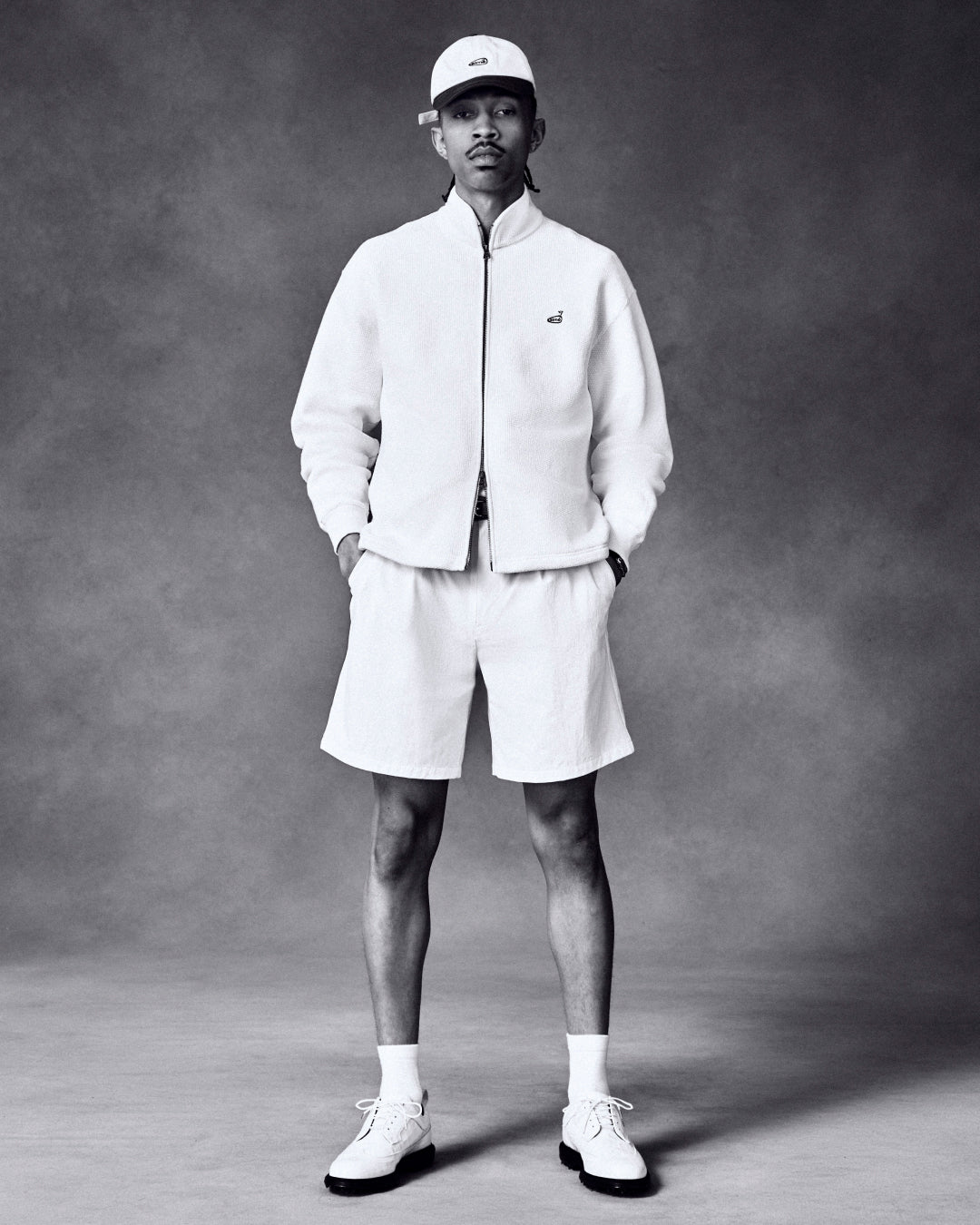 Man wears ALD Golf Piqué Full Zip Sweater, shorts, cap, socks, and shoes, hands in pockets against plain backdrop.