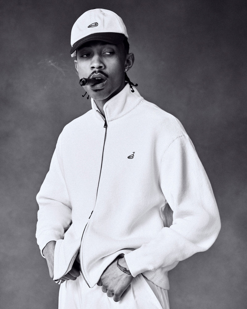 Man wearing ALD Golf Logo Hat and light jacket smokes a cigar, looking sideways against a plain studio backdrop.