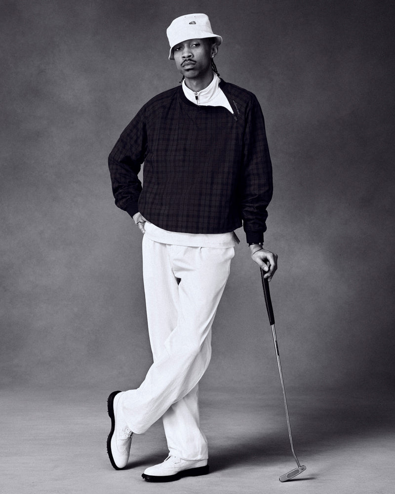 Man wears ALD Golf Cordura Double Pleated Pant, dark sweater, and bucket hat while posing with golf club in studio.