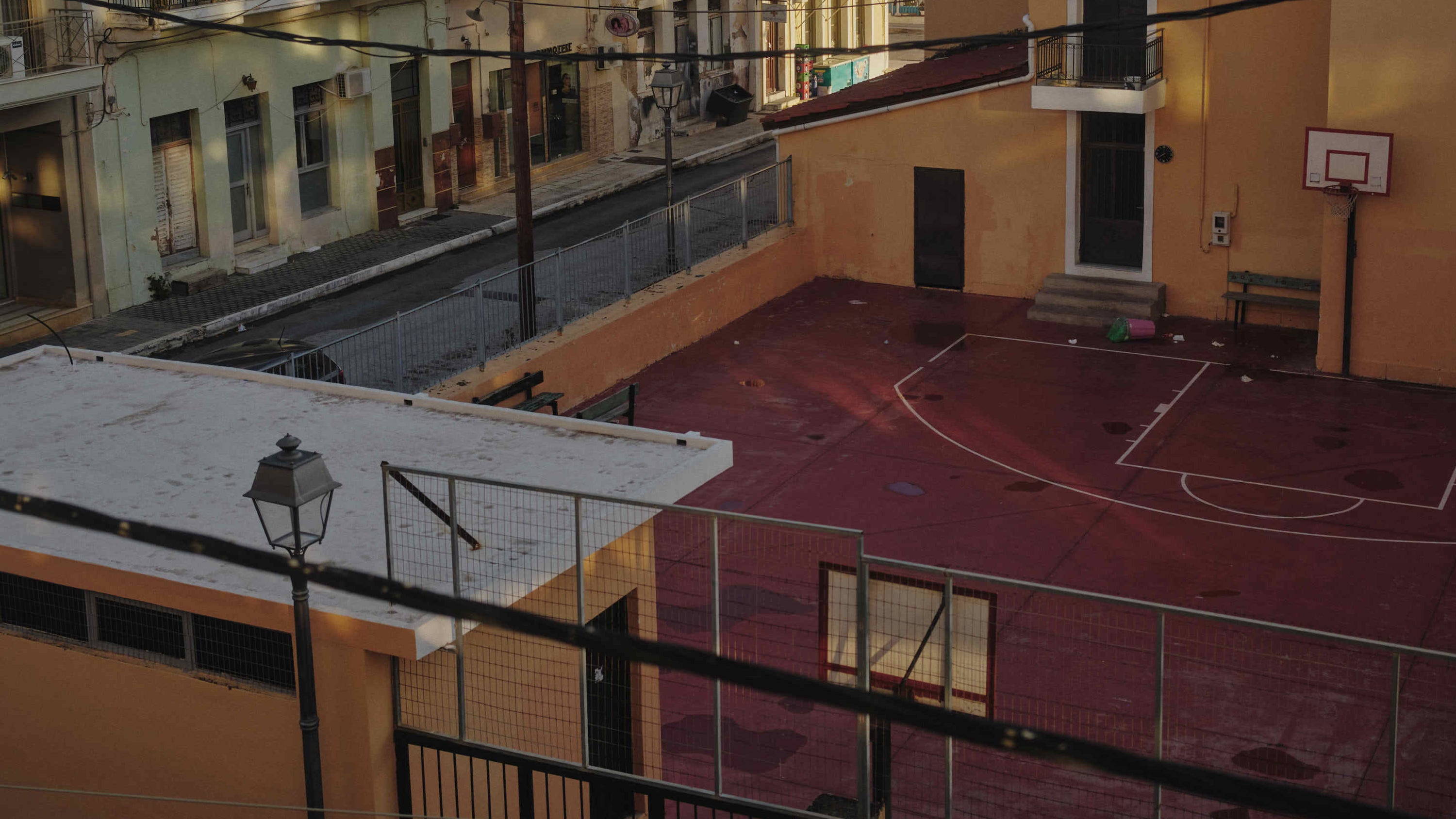 An elevated view of an outdoor basketball court with a red-pink painted surface, surrounded by low buildings with yellow-orange walls and a black metal fence in the foreground.