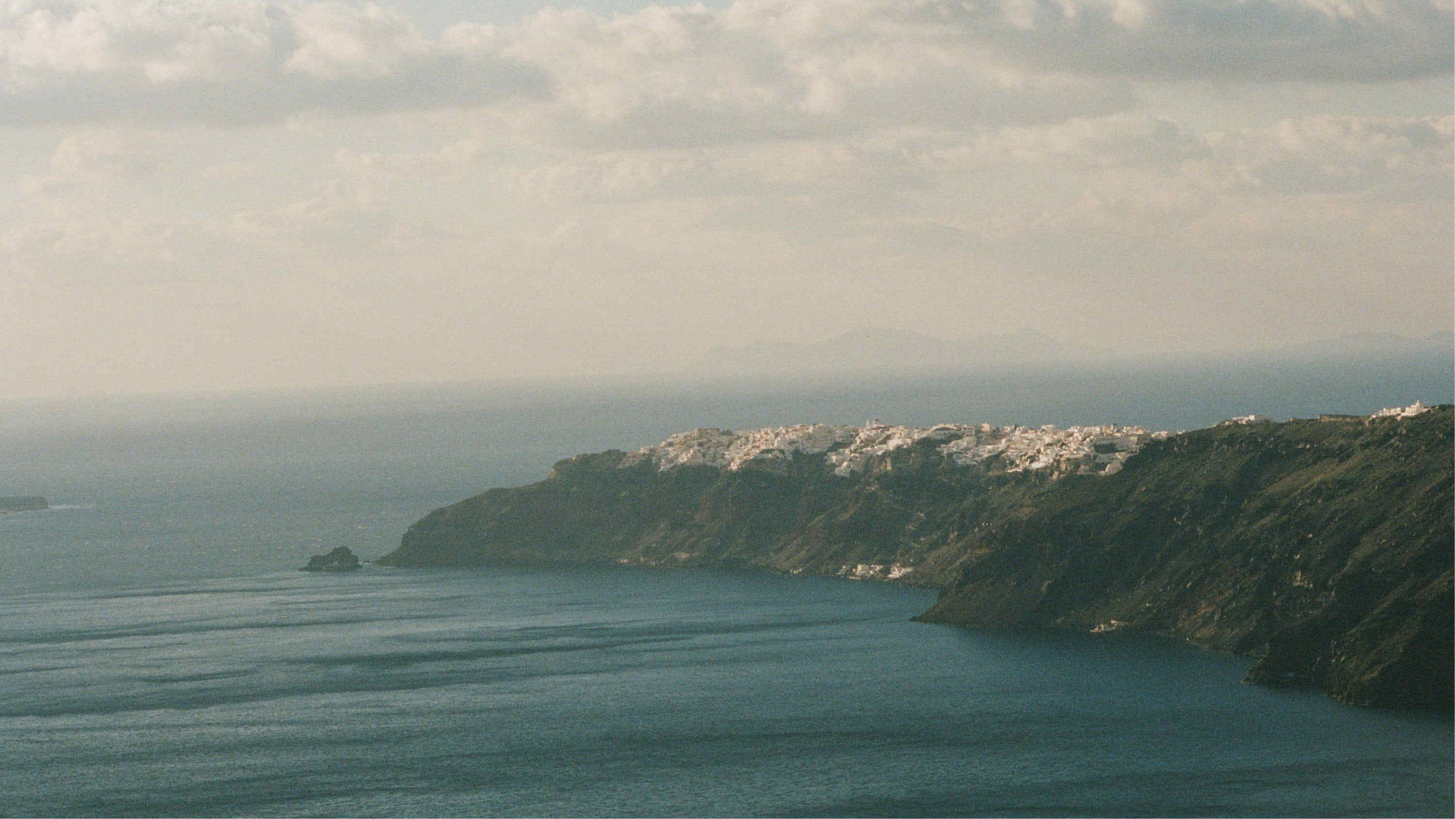 A scenic coastal landscape with deep blue water in the foreground, a dark green cliffside on the right, and a cluster of white buildings on the cliff top under a cloudy sky.