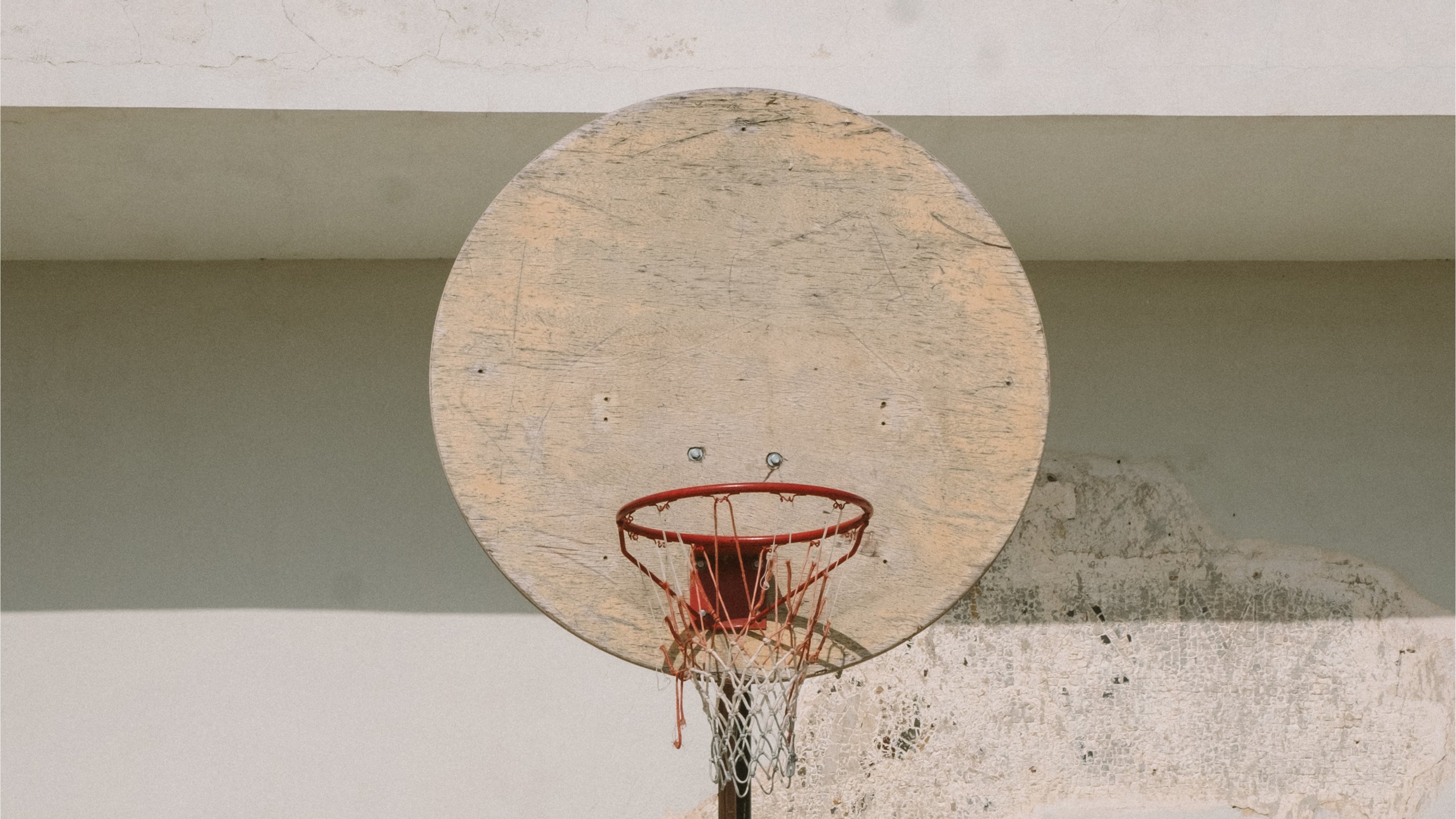 A close-up view of a weathered circular basketball backboard with a red rim and net, mounted against a concrete wall.
