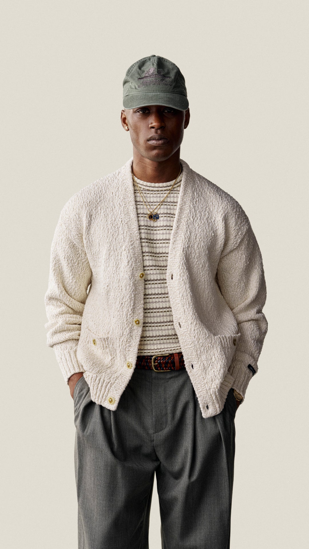 A man wearing a cream textured knit button-up cardigan over a striped knit shirt, paired with dark gray trousers, a brown belt, and an olive green baseball cap, standing against a solid light beige background.