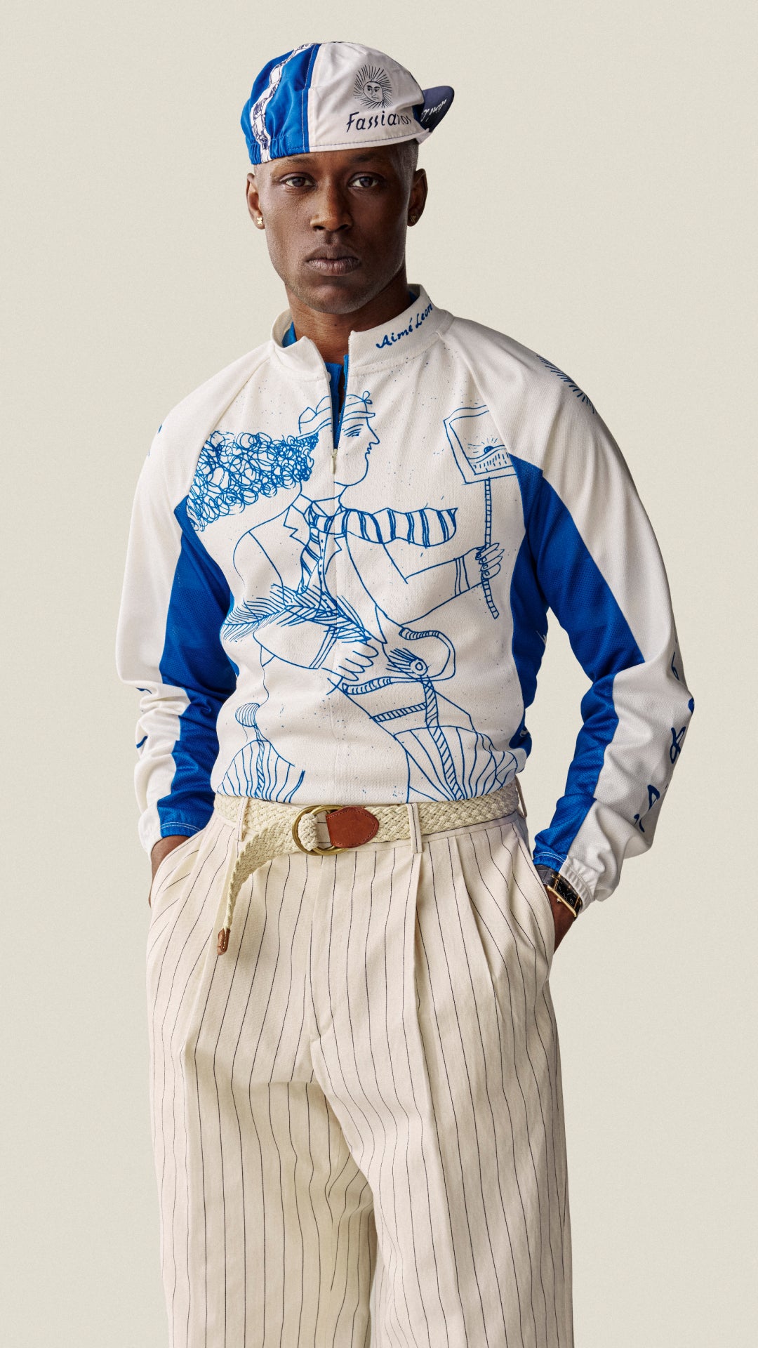 Man wearing a white and blue jacket with graphic design, beige pants, and a cap on a plain background
