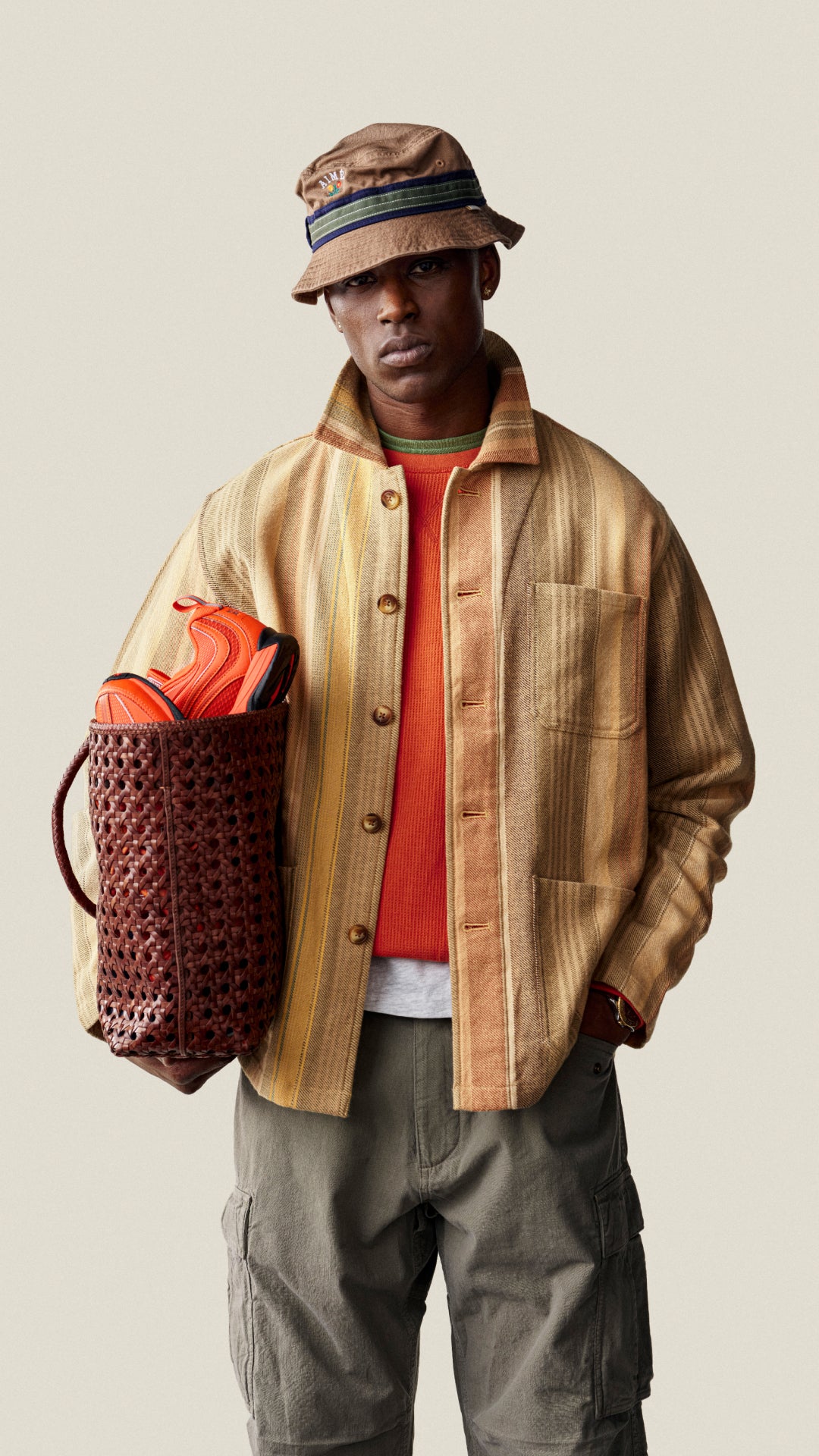 A model wearing a tan and yellow striped button-up shirt jacket, an orange t-shirt, olive cargo pants, and a brown bucket hat, holding a brown woven basket bag with orange shoes inside, against a solid light beige background.