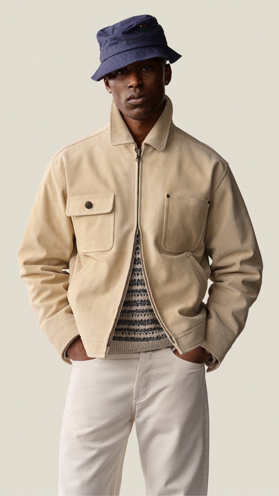 Man wearing a beige jacket, white pants, and a blue bucket hat on a plain background