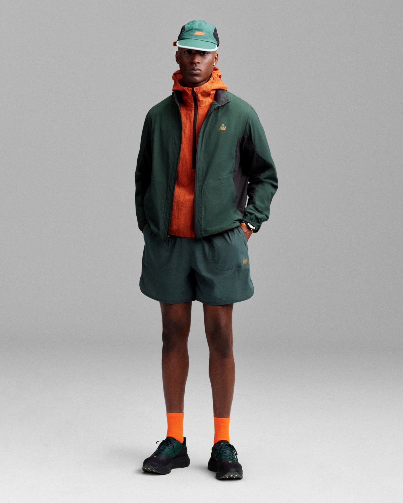 A person in the ALD / New Balance Performance Nylon Jacket, shorts, cap, hoodie, and socks stands by a grey background.