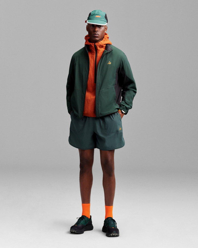 A person in a green jacket, orange hoodie, green shorts, orange socks, black shoes, and an ALD/New Balance Mesh Sport Hat stands facing forward.