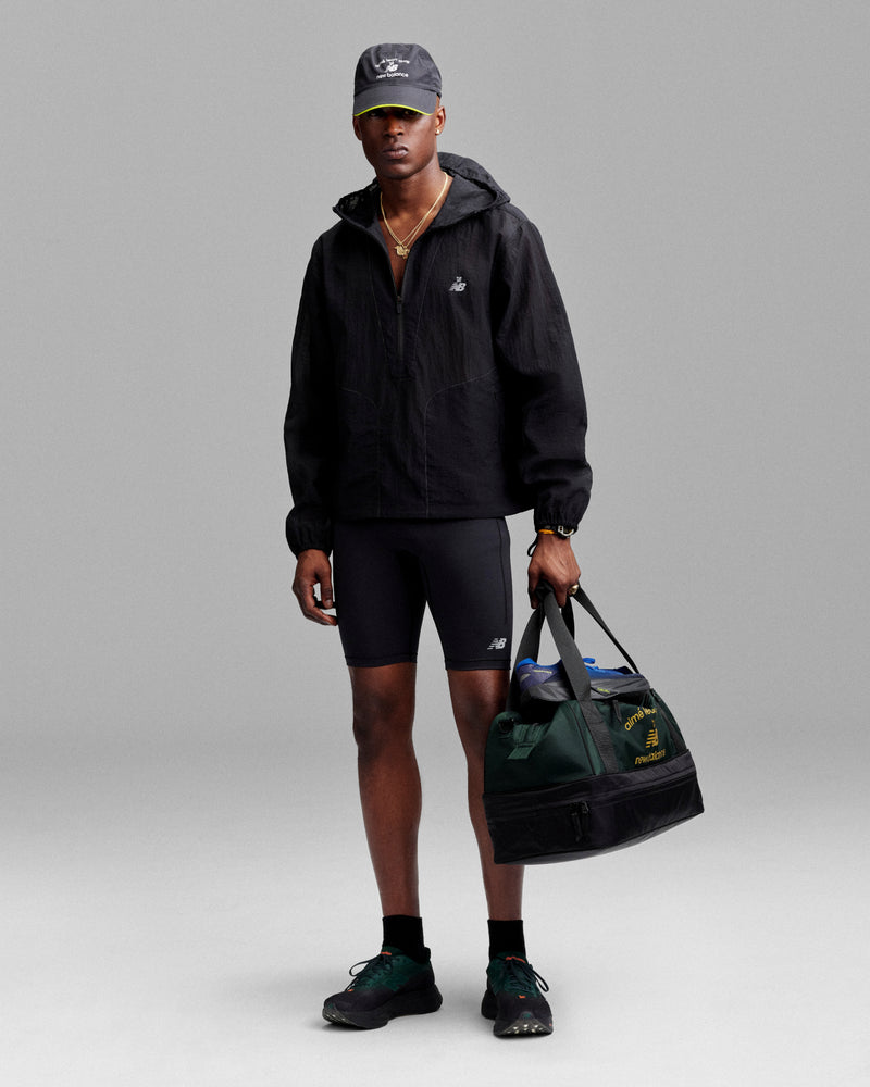 Man in black jacket, bike shorts, and cap holds an ALD / New Balance Travel Duffle Bag against a gray background.