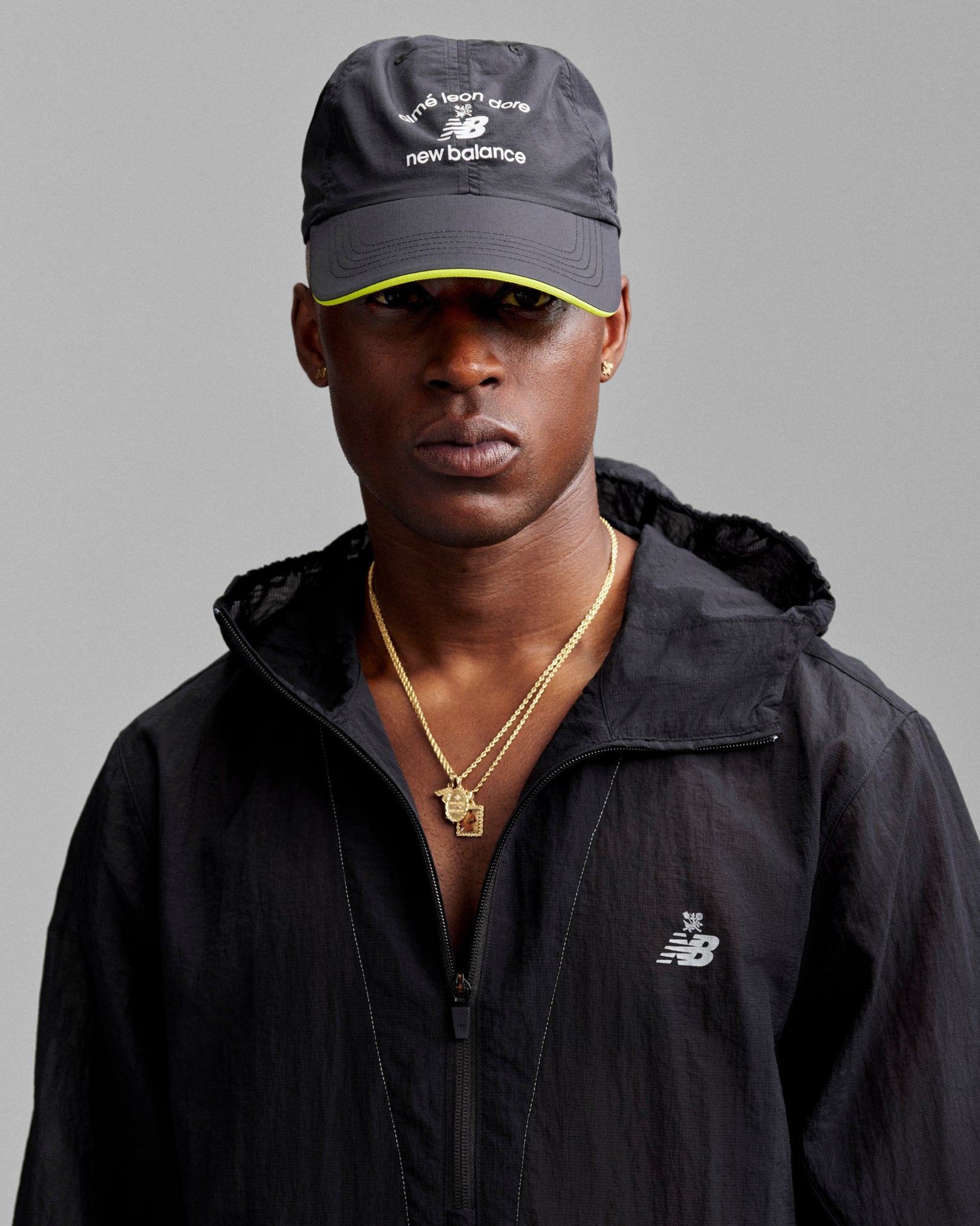 A man in a black jacket and ALD / New Balance Running Hat, with gold necklaces, poses against a gray background.