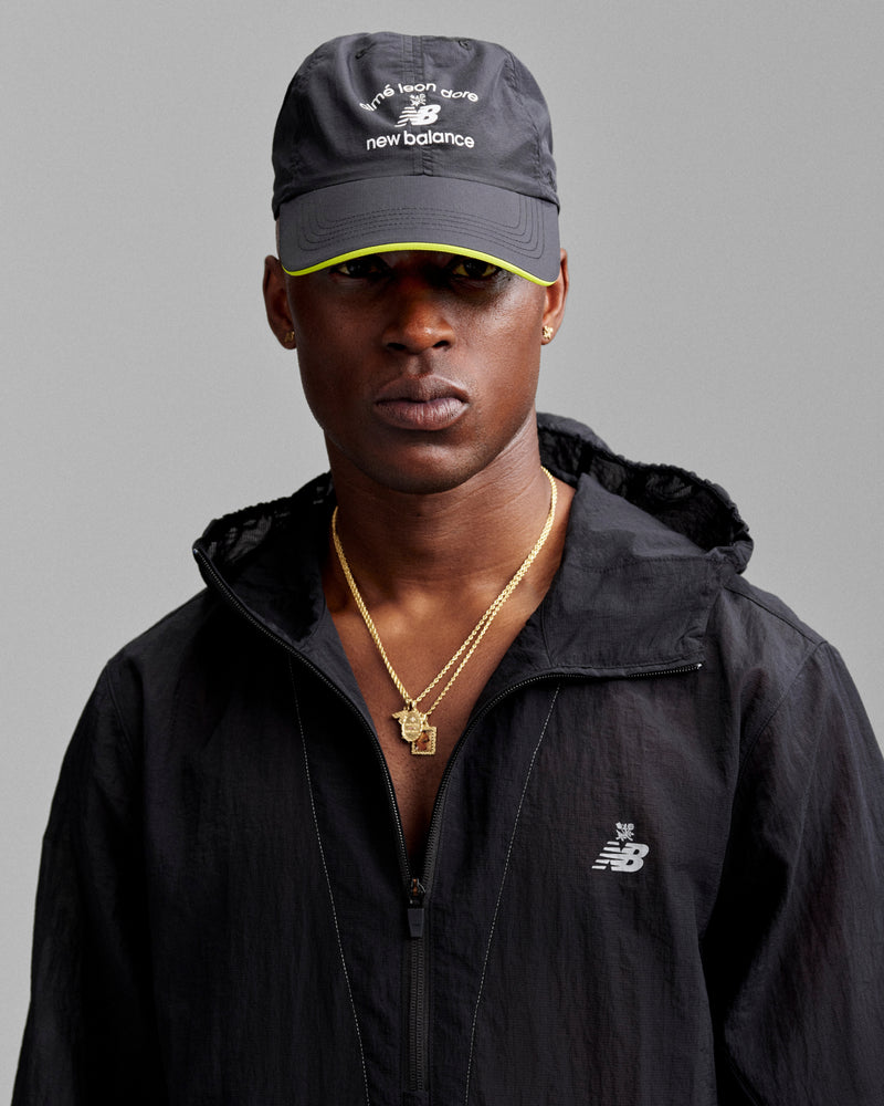 A man in a black jacket and ALD / New Balance Running Hat, with gold necklaces, poses against a gray background.