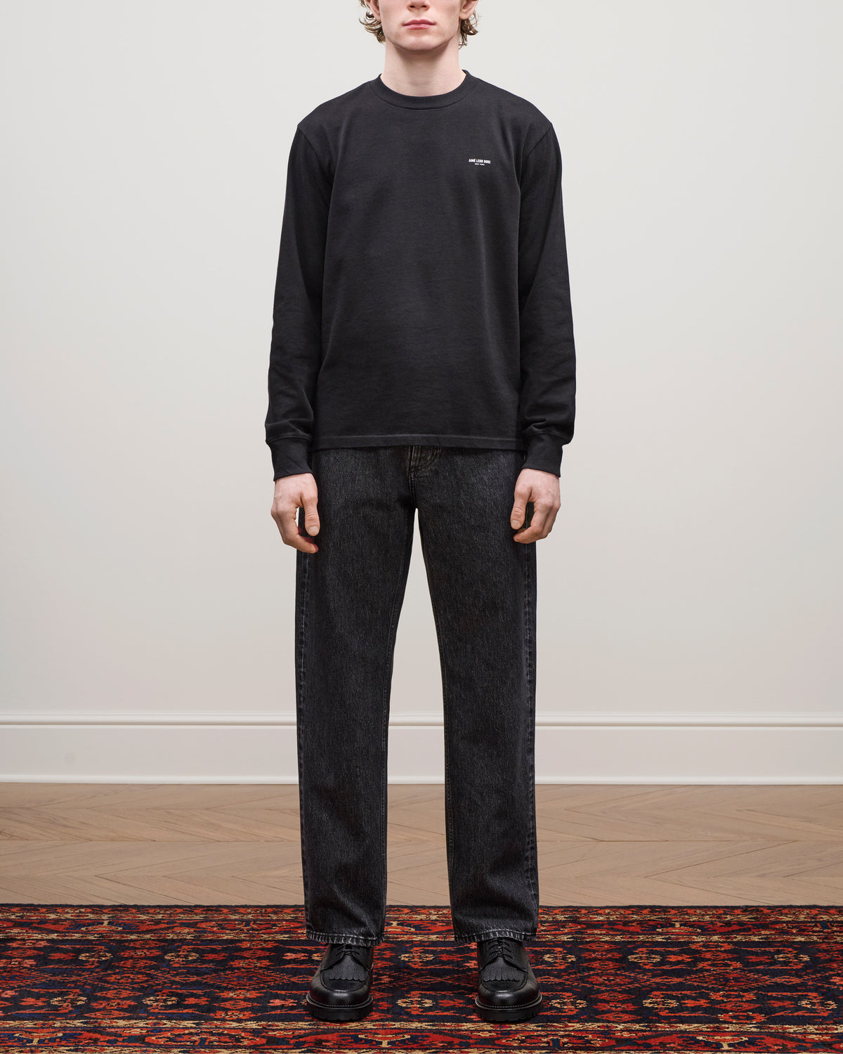 Person standing on a patterned rug wearing the Long-Sleeve Micro Logo Tee, black jeans, and black shoes.