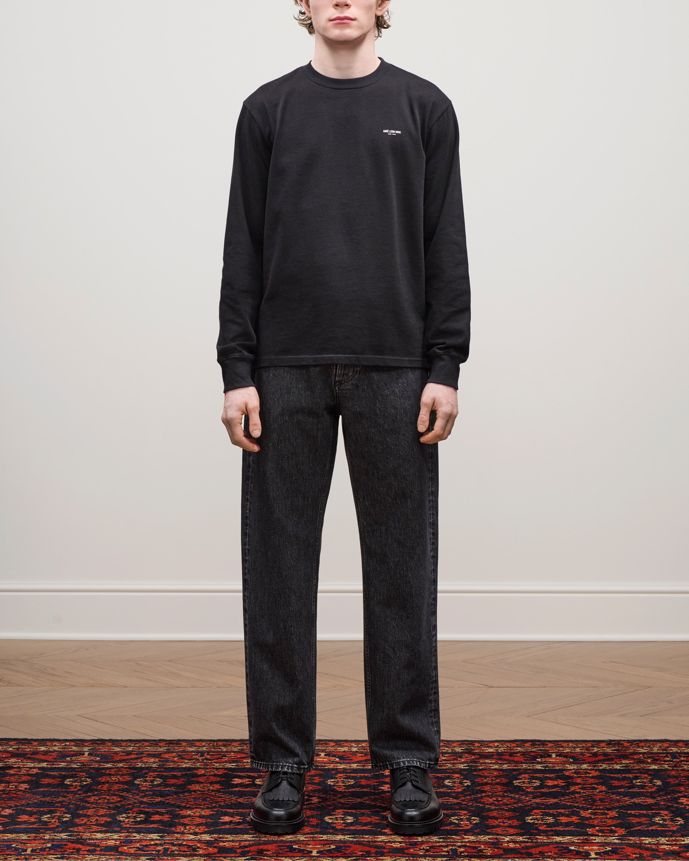 Person standing on a patterned rug wearing the Long-Sleeve Micro Logo Tee, black jeans, and black shoes.