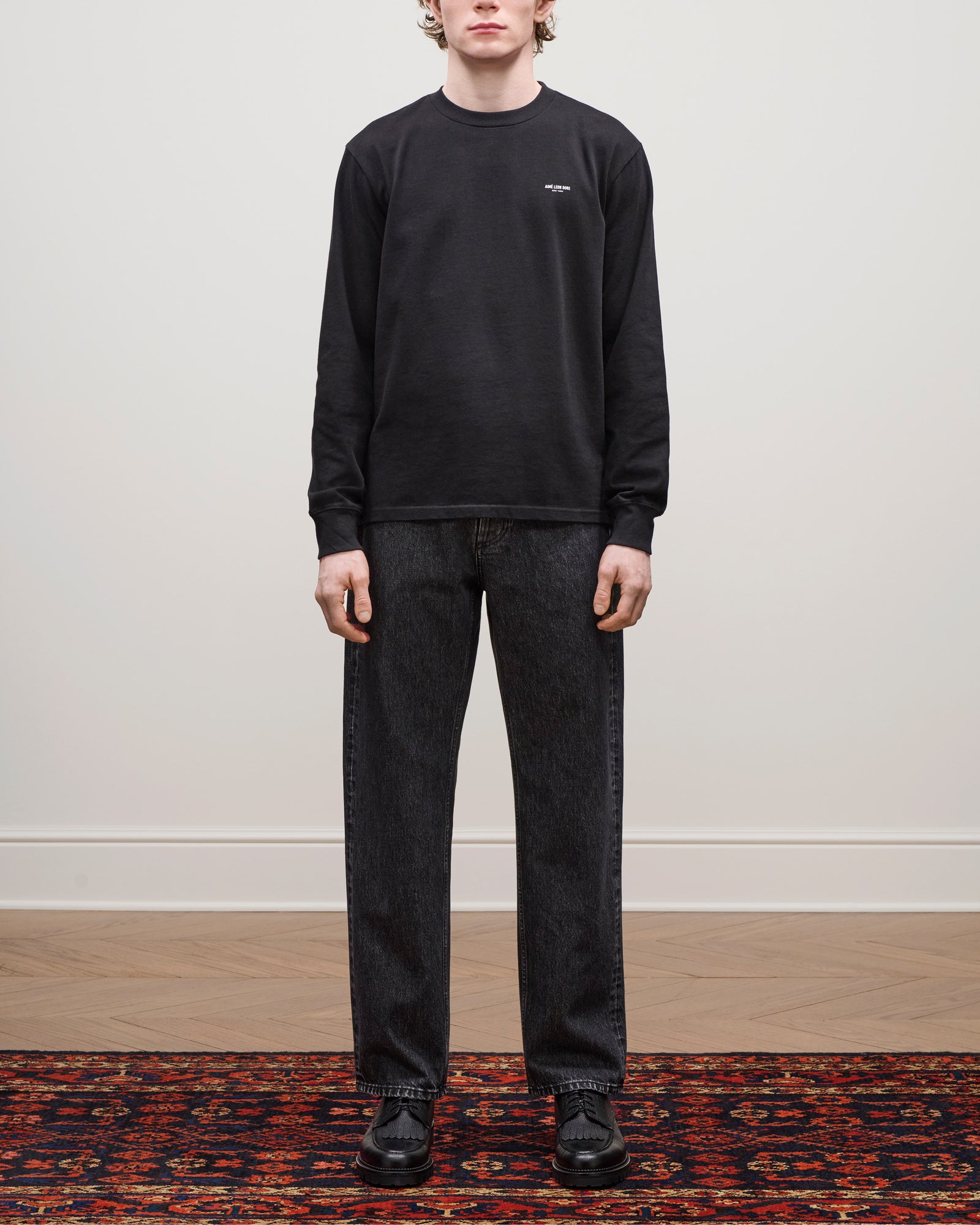 A person stands indoors on a patterned rug wearing Relaxed Fit 5-Pocket Denim Pant, a black sweatshirt, and black shoes.