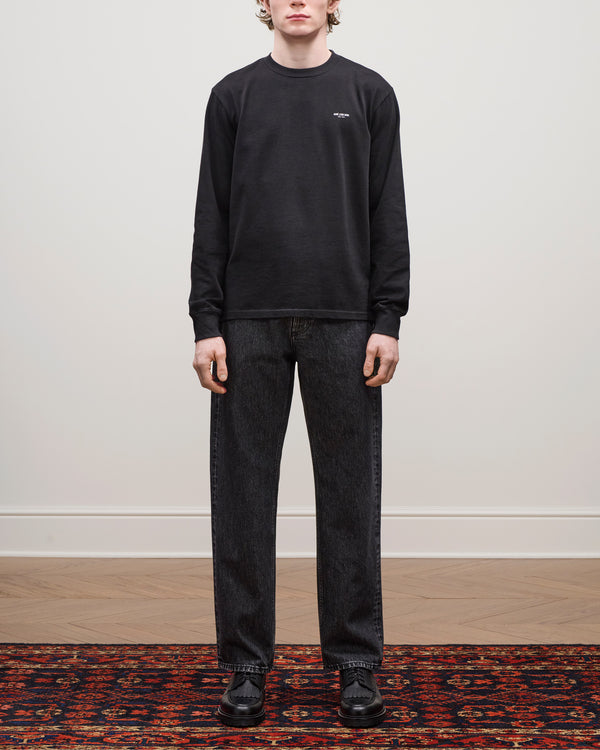 A person stands indoors on a patterned rug wearing Relaxed Fit 5-Pocket Denim Pant, a black sweatshirt, and black shoes.