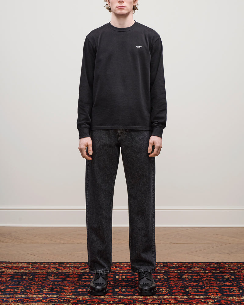 A person stands indoors on a patterned rug wearing Relaxed Fit 5-Pocket Denim Pant, a black sweatshirt, and black shoes.