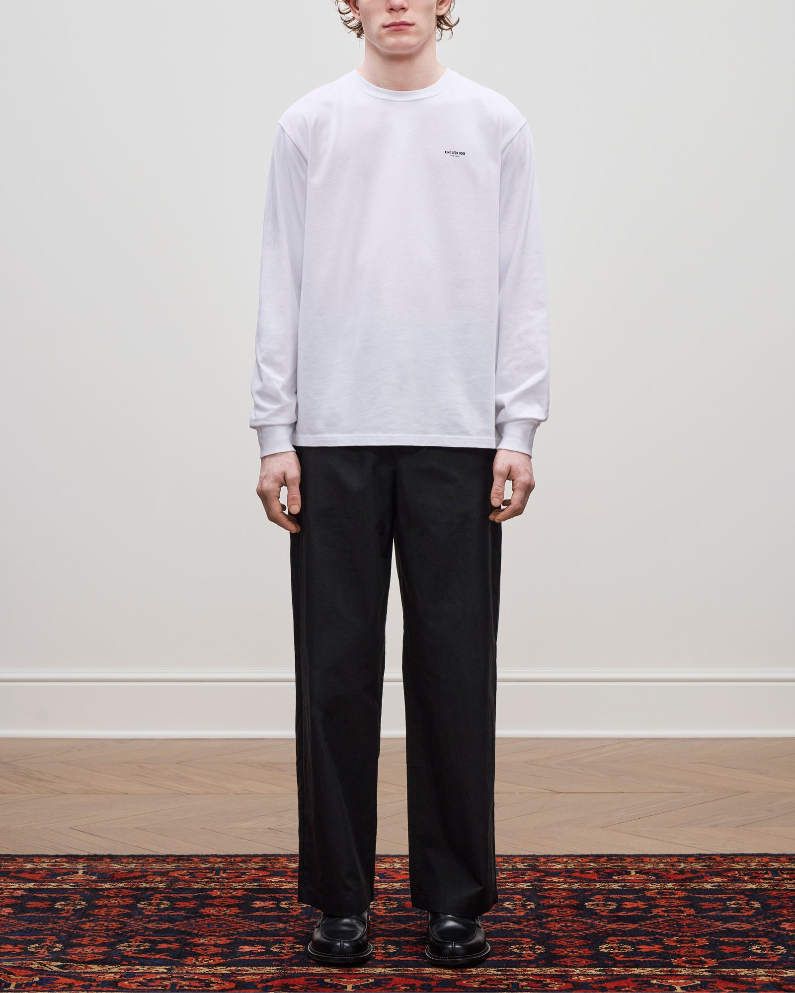 Person wearing a Long-Sleeve Micro Logo Tee, black pants, and black shoes, standing on a patterned rug indoors.